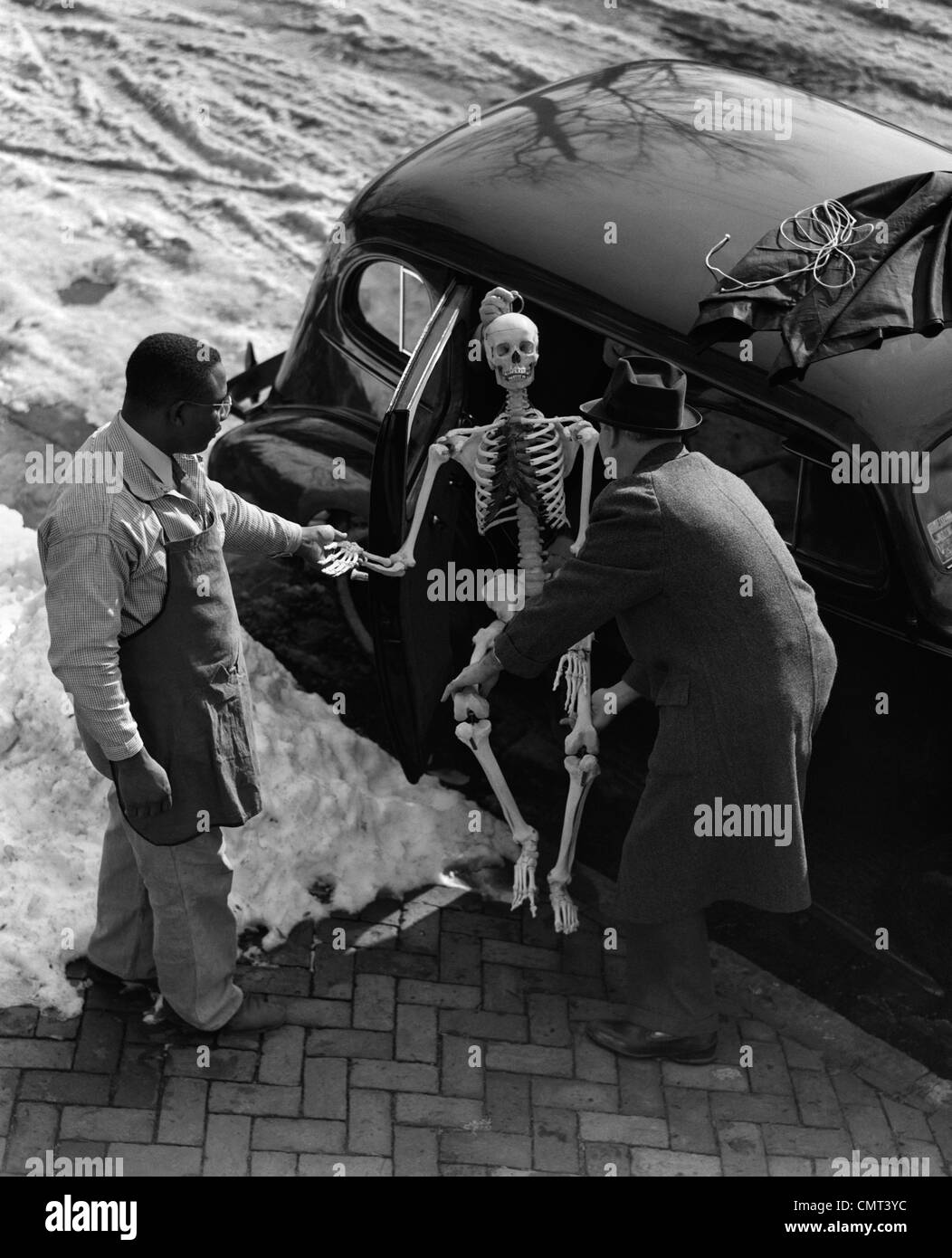 1930s TWO MEN TAKING SKELETON OUT OF BACK SEAT OF CAR OUTDOOR WINTER Stock Photo