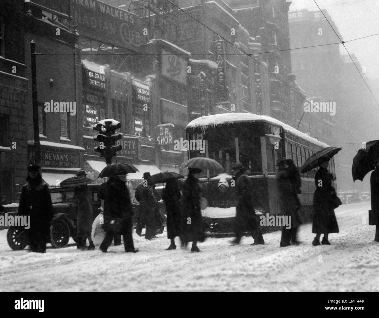 1920s 1930s CROWD CARRYING UMBRELLAS CROSSING STREET IN FRONT OF STREET CAR TROLLEY DURING SNOWSTORM Stock Photo