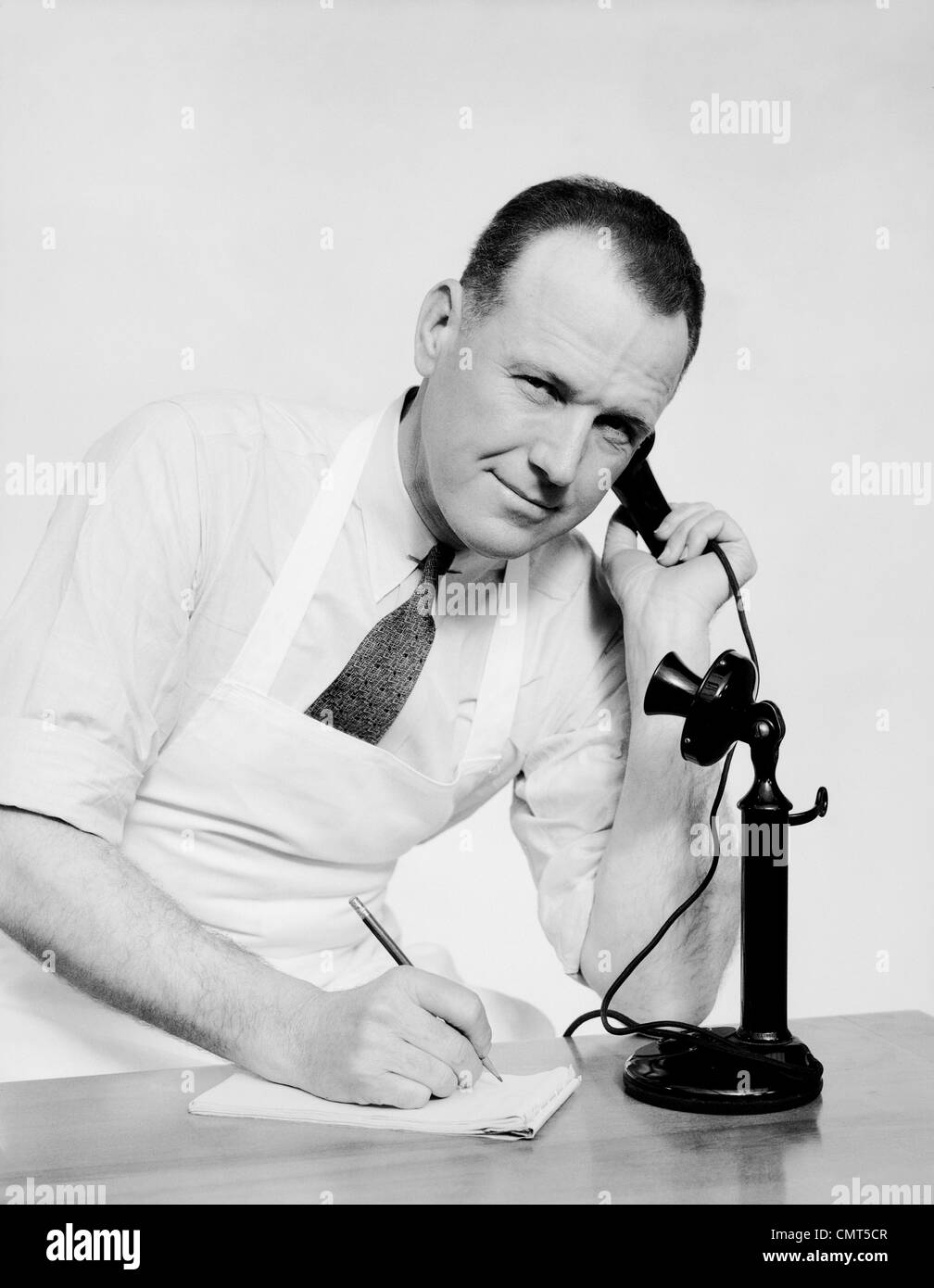 1930s PORTRAIT MAN CLERK WEARING APRON TALKING ON OLD BLACK CANDLESTICK TELEPHONE WRITING ON PAD WITH PENCIL LOOKING AT CAMERA Stock Photo