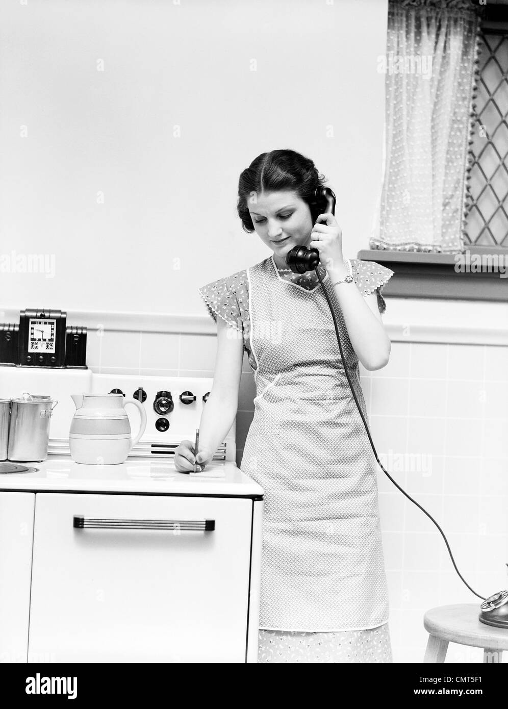 1930s WOMAN HOUSEWIFE STANDING KITCHEN BY STOVE TALKING ON TELEPHONE WRITING ON PAD WEARING APRON Stock Photo