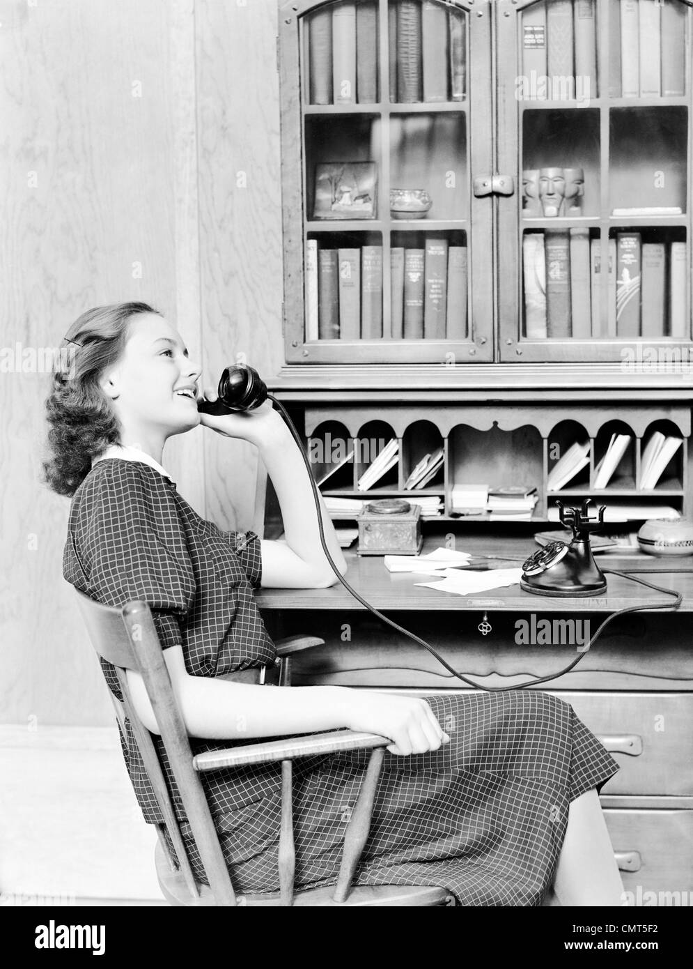1930s 1940s TEENAGE GIRL SITTING AT DESK TALKING ON OLD BLACK ROTARY DIAL PHONE Stock Photo