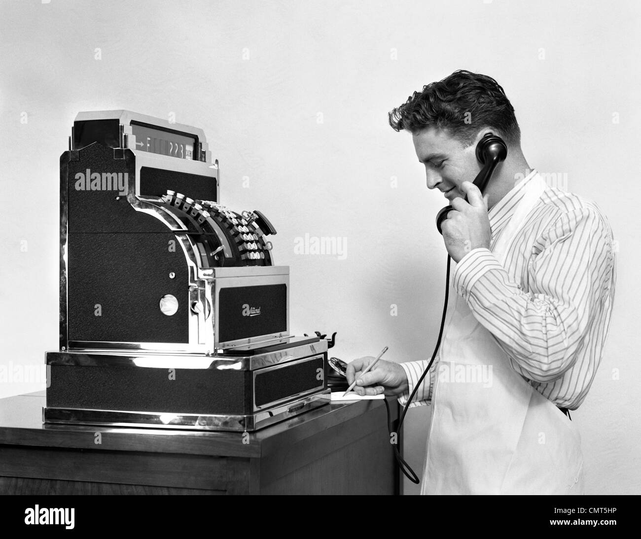 1930s 1940s MAN IN APRON NEXT TO LARGE CASH REGISTER TALKING ON PHONE TAKING NOTES SMILING Stock Photo