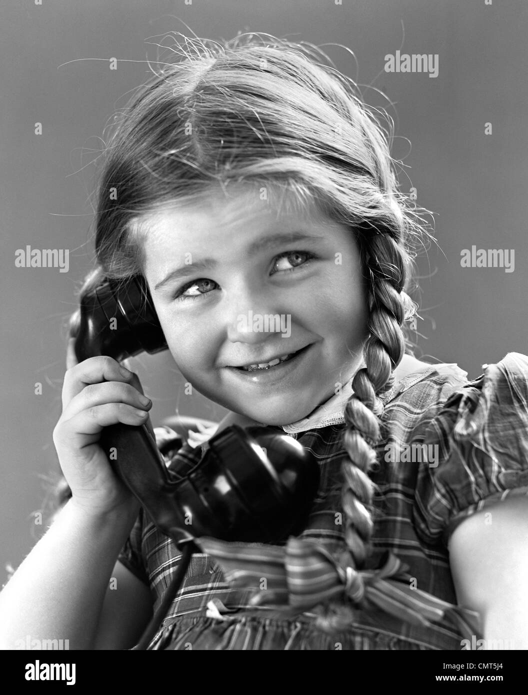 1930s 1940s PORTRAIT OF SMILING LITTLE GIRL WITH LONG PIGTAILS TALKING ON TELEPHONE Stock Photo