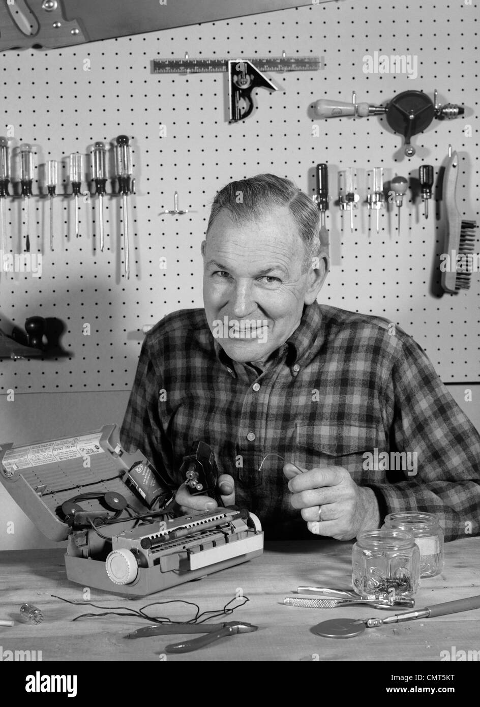 1960s MAN REPAIRING PORTABLE RADIO IN HOME WORKSHOP SMILING LOOKING AT CAMERA INDOOR Stock Photo
