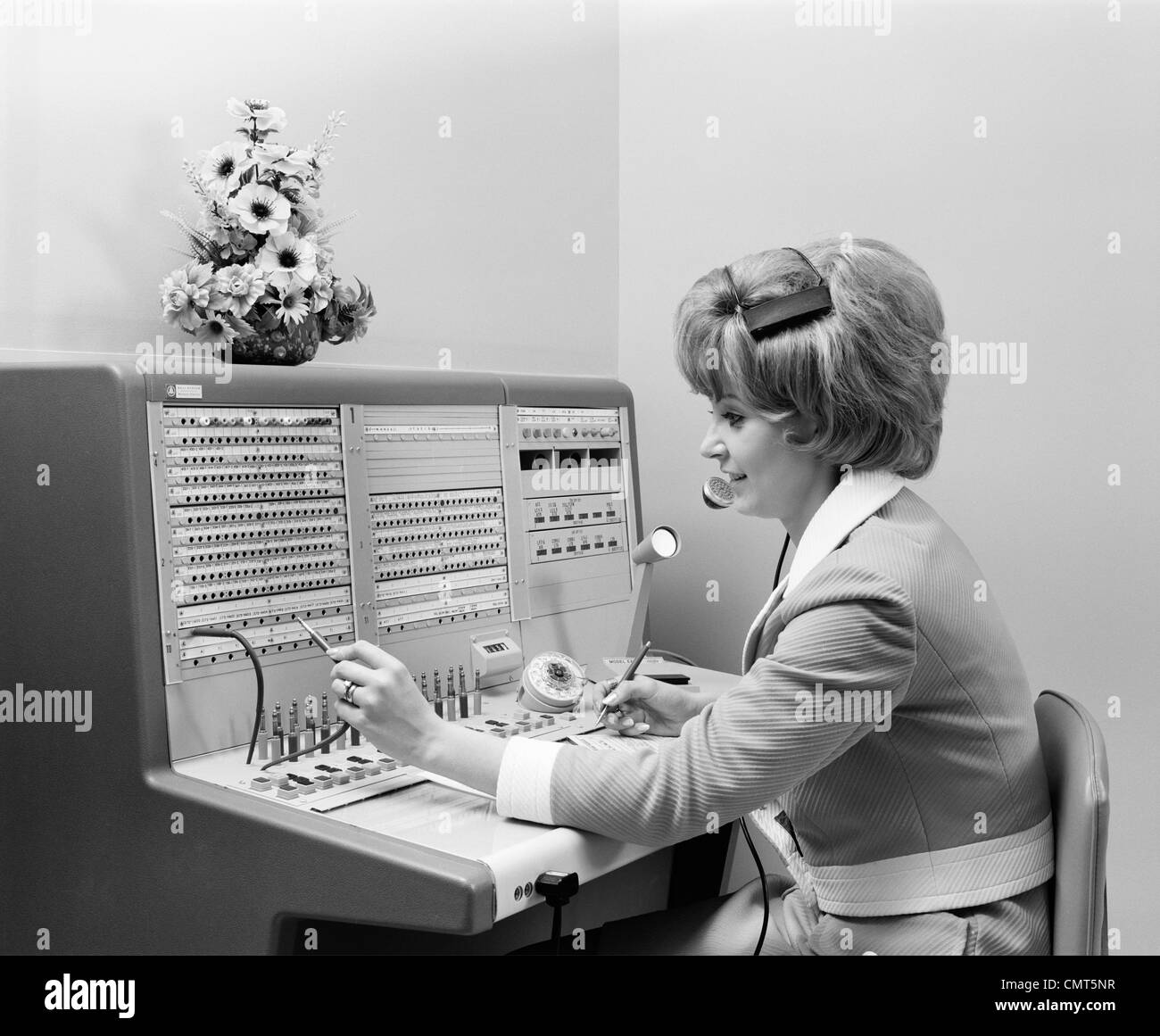 1970s SMILING WOMAN OPERATOR RECEPTIONIST WORKING ANSWERING OFFICE TELEPHONE SWITCHBOARD WEARING HEADSET INDOOR Stock Photo