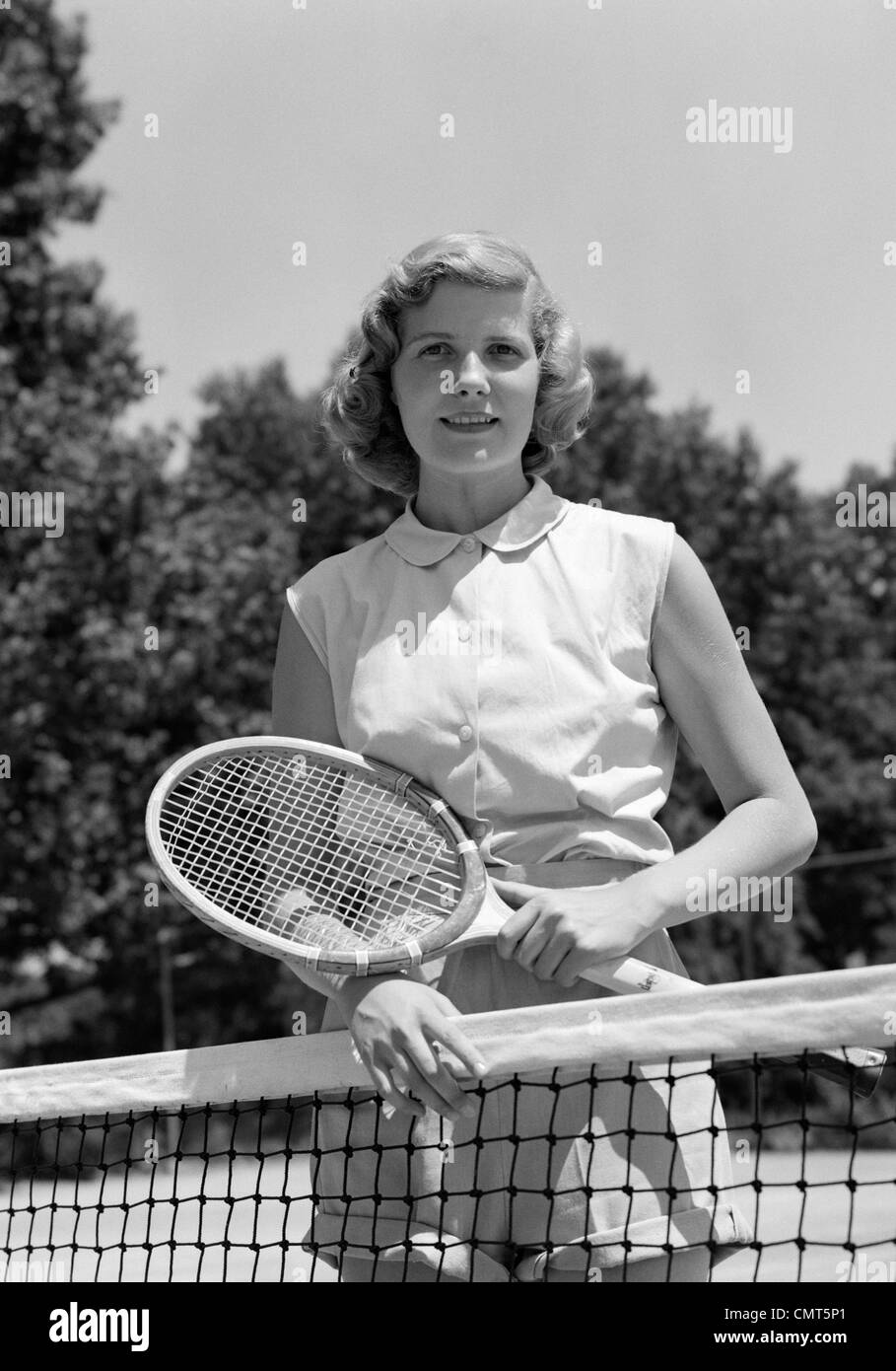 1950s PORTRAIT WOMAN HOLDING TENNIS RACQUET STANDING BEHIND NET OUTDOOR LOOKING AT CAMERA Stock Photo