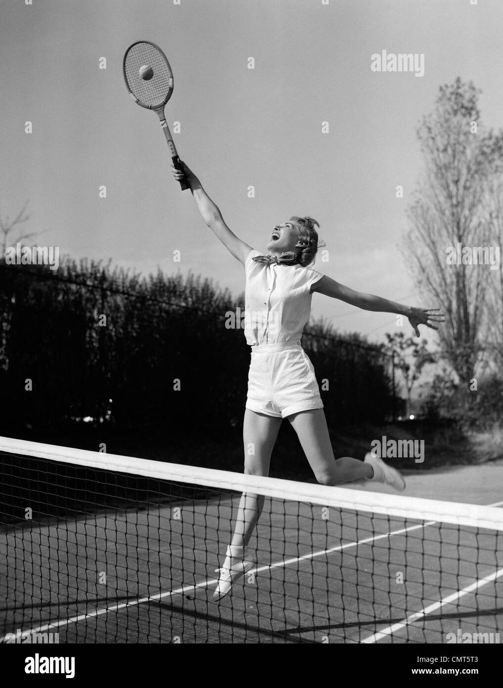 1950s WOMAN JUMPING TO HIT TENNIS BALL Stock Photo