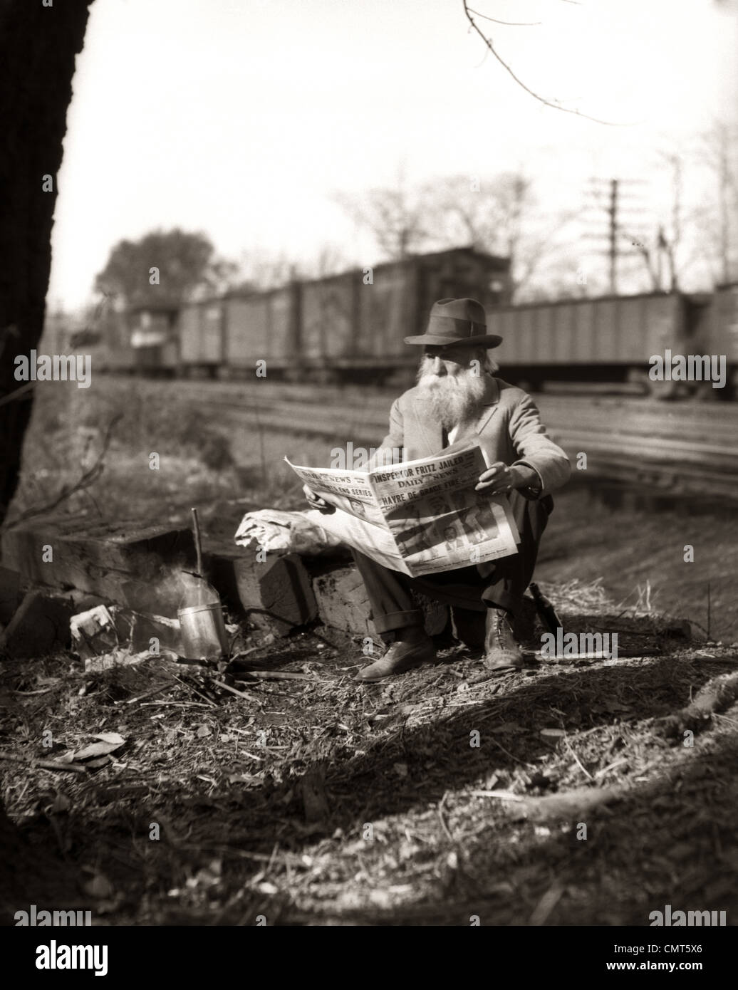 1930s HOBO SITTING BY RAILROAD TRACK READING NEWSPAPER DURING GREAT DEPRESSION Stock Photo