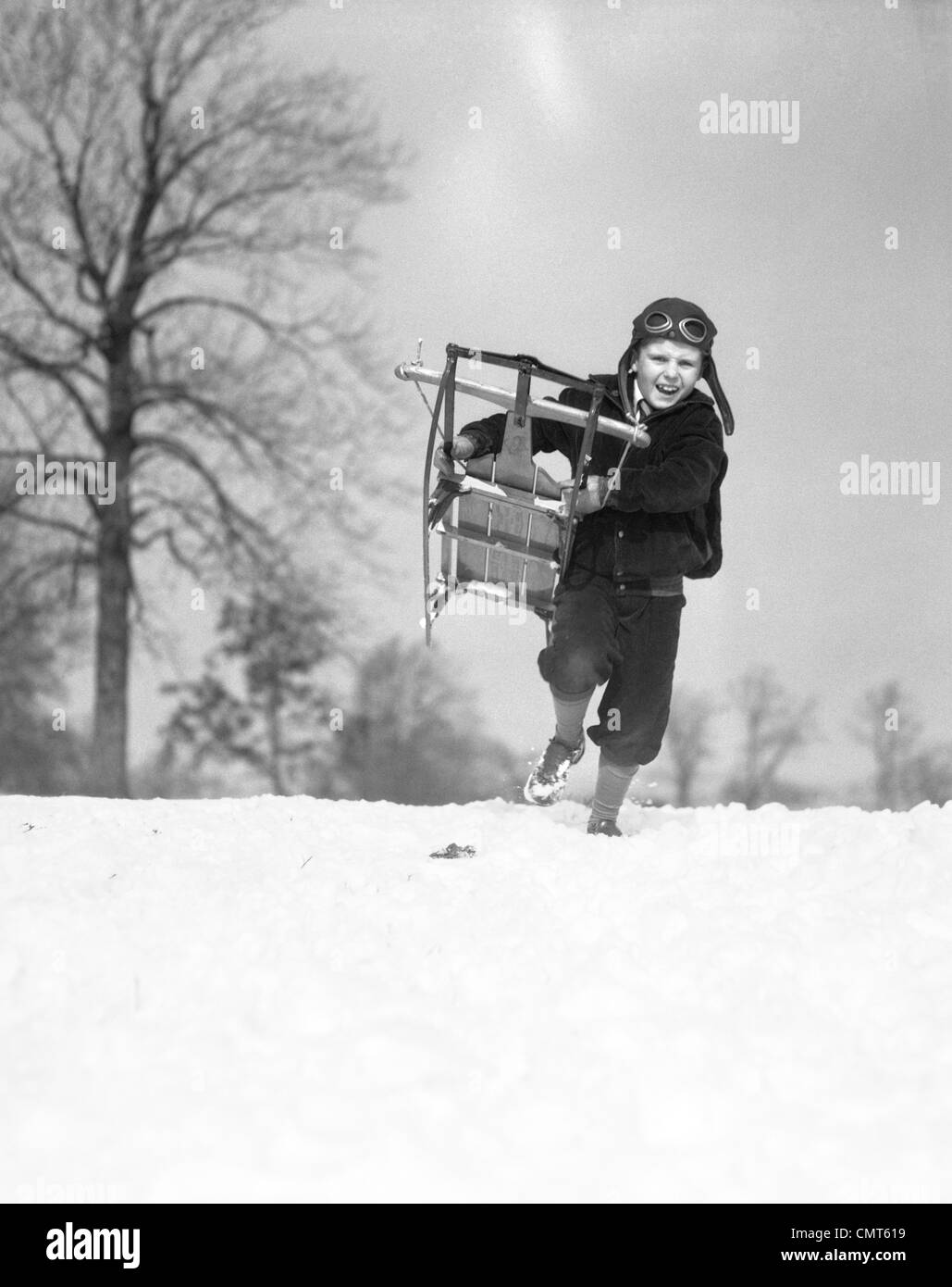 1930s BOY WEARING AVIATOR CAP RUNNING THROUGH SNOW WITH SLED BELLY FLOPPING Stock Photo