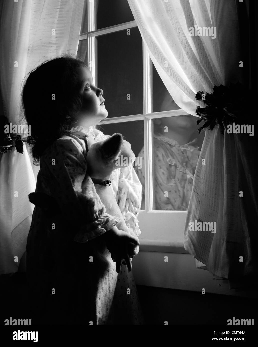 1950s GIRL IN FLANNEL NIGHTGOWN HOLDING STUFFED ANIMAL LOOKING OUT WINDOW AT NIGHT WITH CURTAINS HELD BACK BY HOLLY Stock Photo