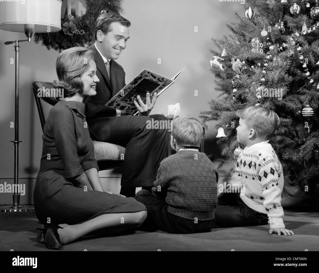 1960s FAMILY FATHER MOTHER TWO SONS SMILING SITTING BY CHRISTMAS TREE IN LIVING ROOM READING A BOOK Stock Photo