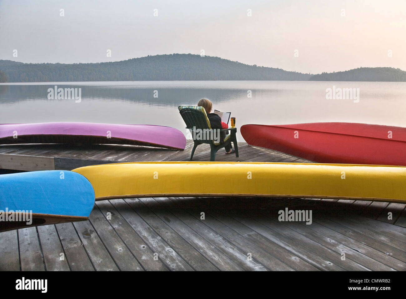 Woman reading on cottage dock surrounded by canoes, Smoke Lake, Algonquin Park, Ontario Stock Photo