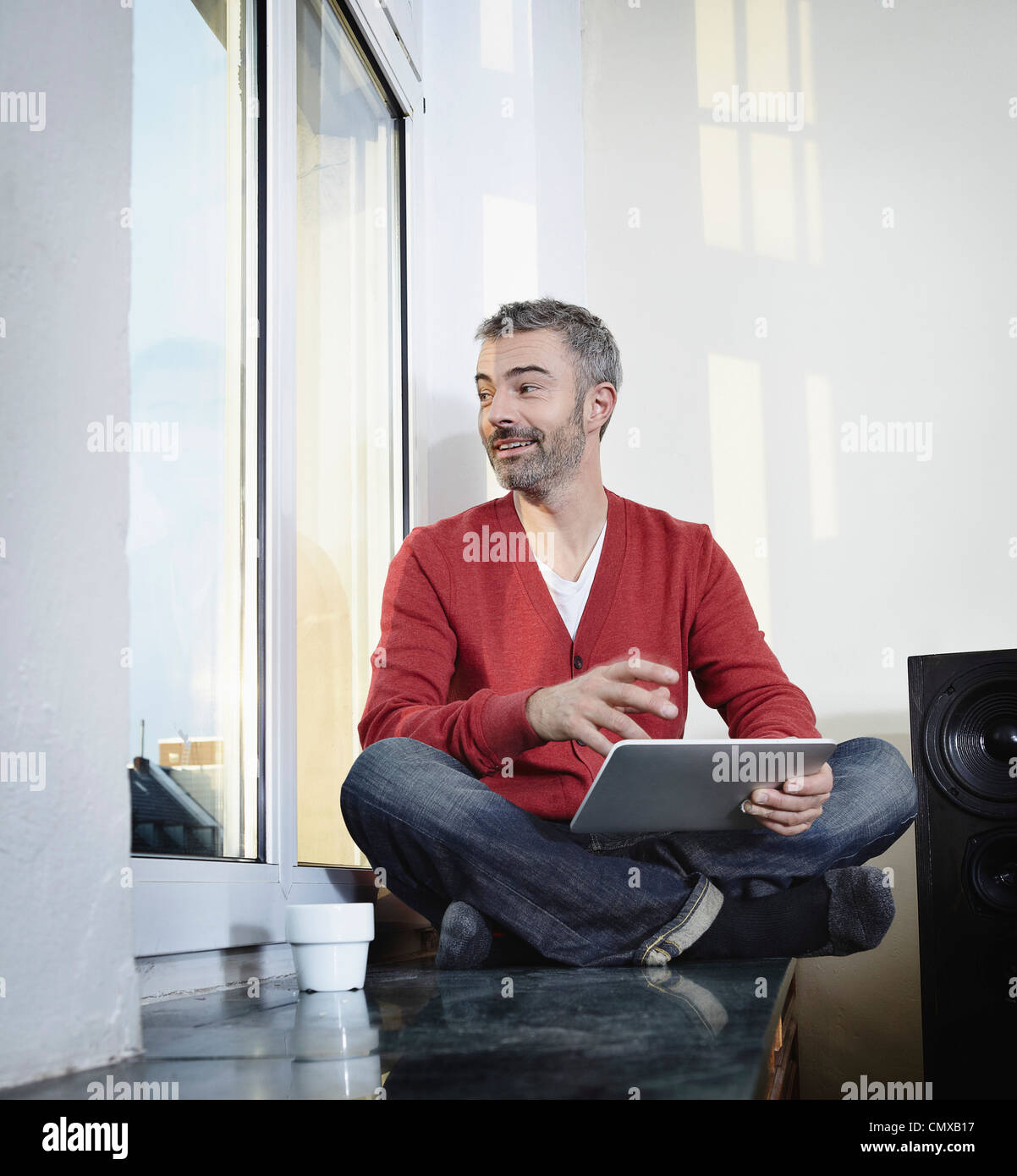 Germany, Cologne, Mature man sitting at window with digital tablet, smiling Stock Photo