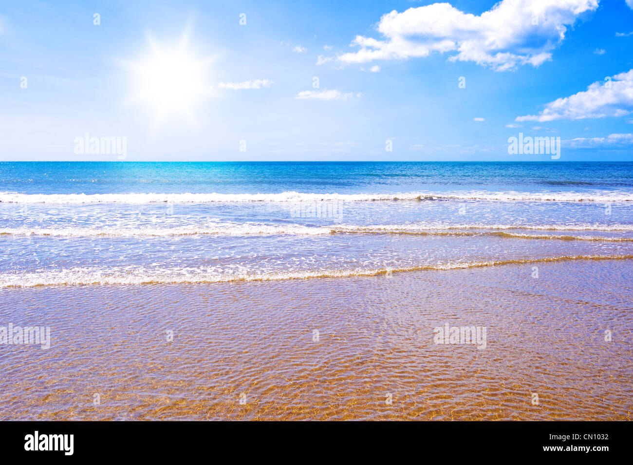 Photo of waves crashing on a golden beach with the sun shining in a blue sky. Stock Photo