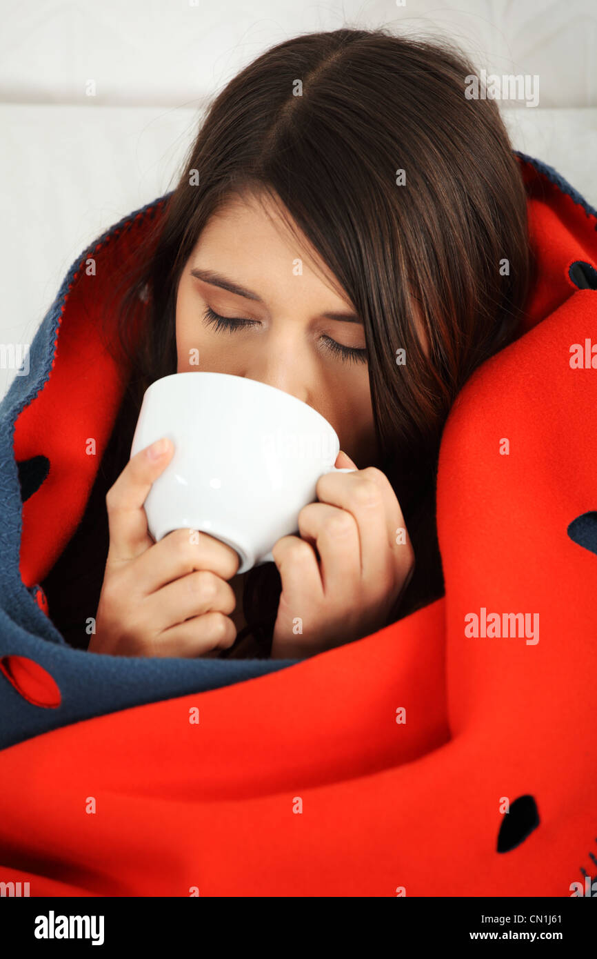 Young woman caught cold, wrapped up in blanket, drinking something hot from cup.  Stock Photo