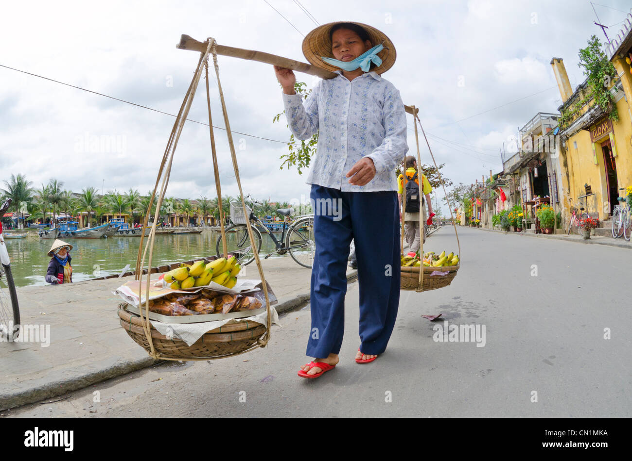 Woman carrying yoke baskets of fruit in street, Hoi An, Vietnam Stock Photo