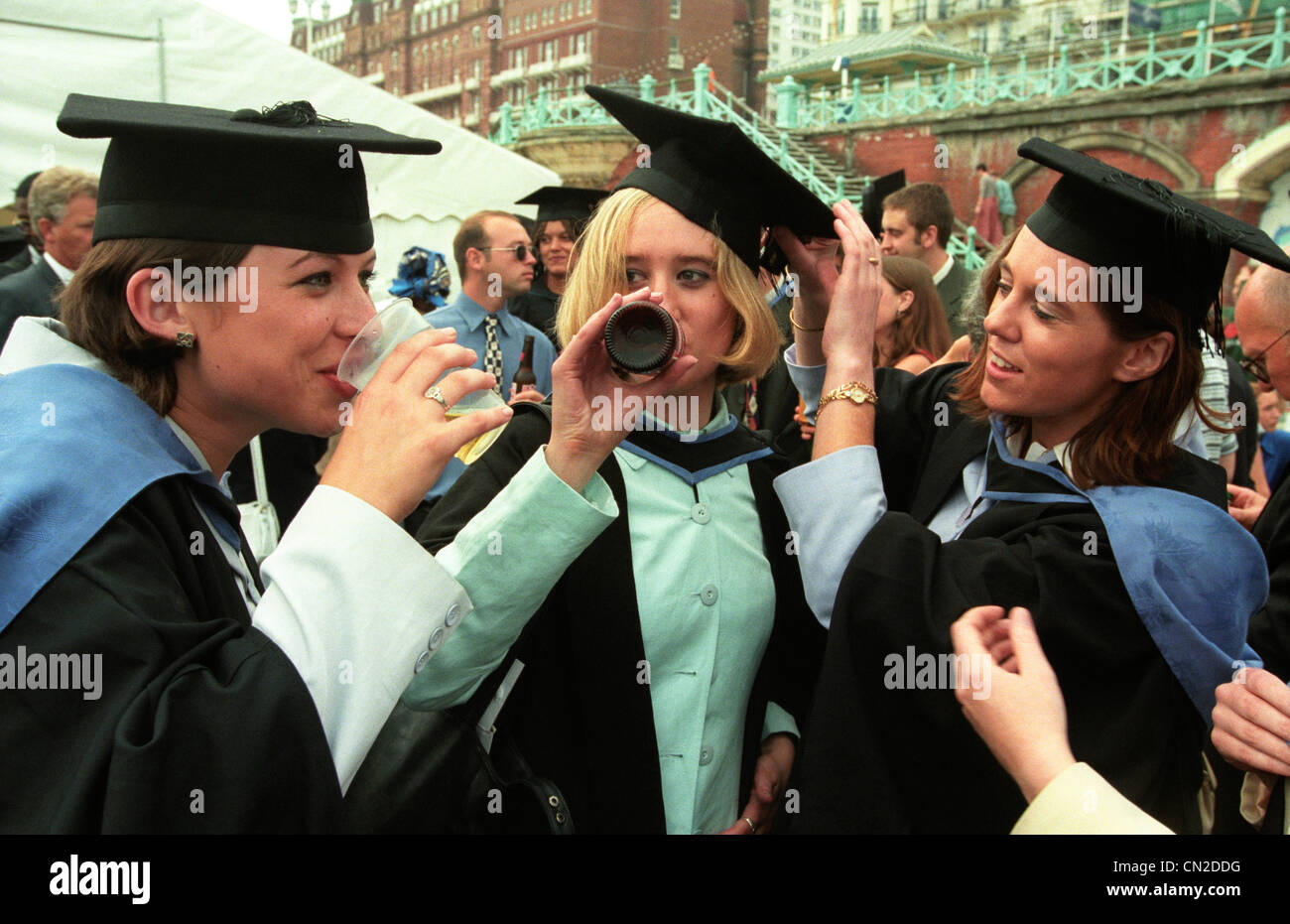A female graduate in gown and mortar board celebrates her degree with a bottle of beer Stock Photo