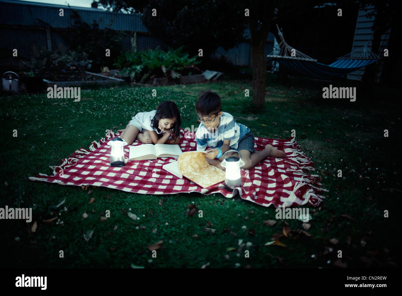 Children read books by lantern light on blanket in garden in evening. Stock Photo