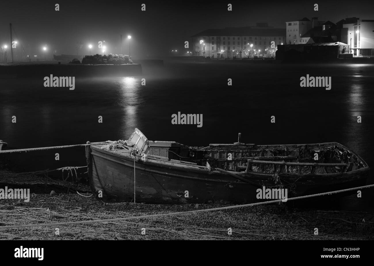 A monochrome shot of an old dilapidated boat tied up in Galway Bay on a misty night with dim lamp lights brightening the horizon Stock Photo