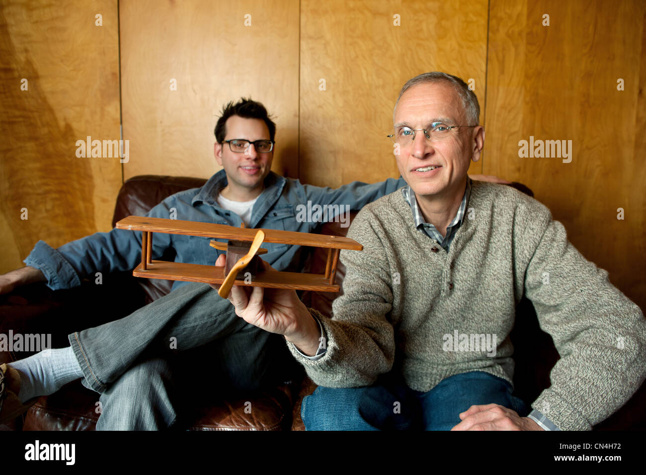 Father holding toy plane with adult son on couch, smiling Stock Photo