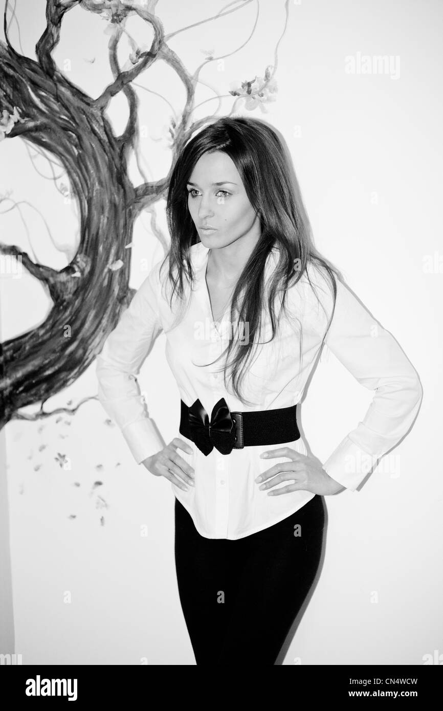 A monochrome portrait of a contemplative woman in a stylish white blouse with a black belt, standing against an artistic backdrop of a painted tree. Stock Photo