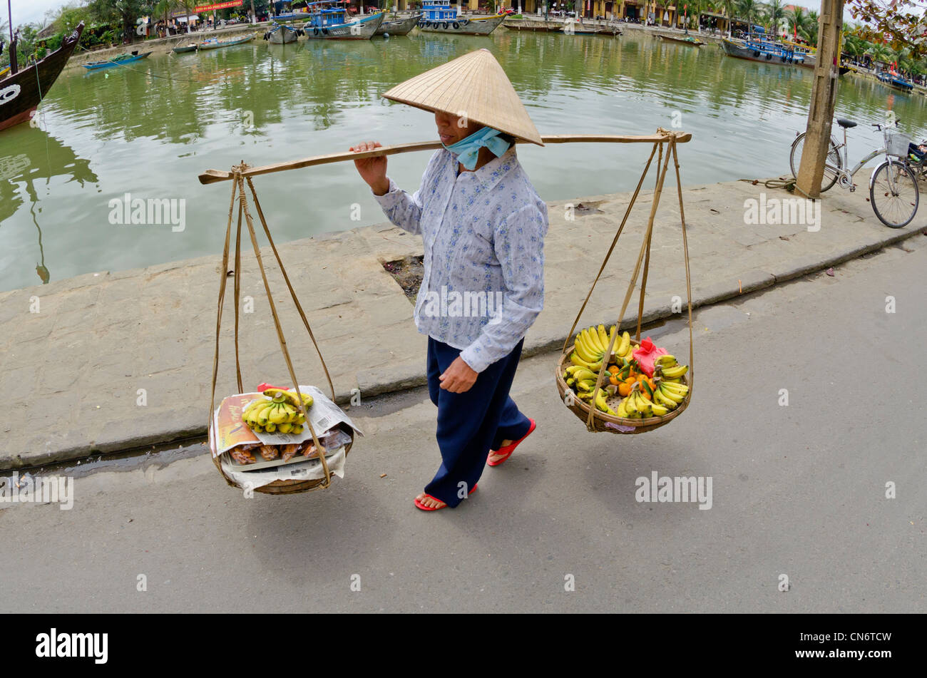 Woman carrying yoke baskets of fruit in street, Hoi An, Vietnam Stock Photo