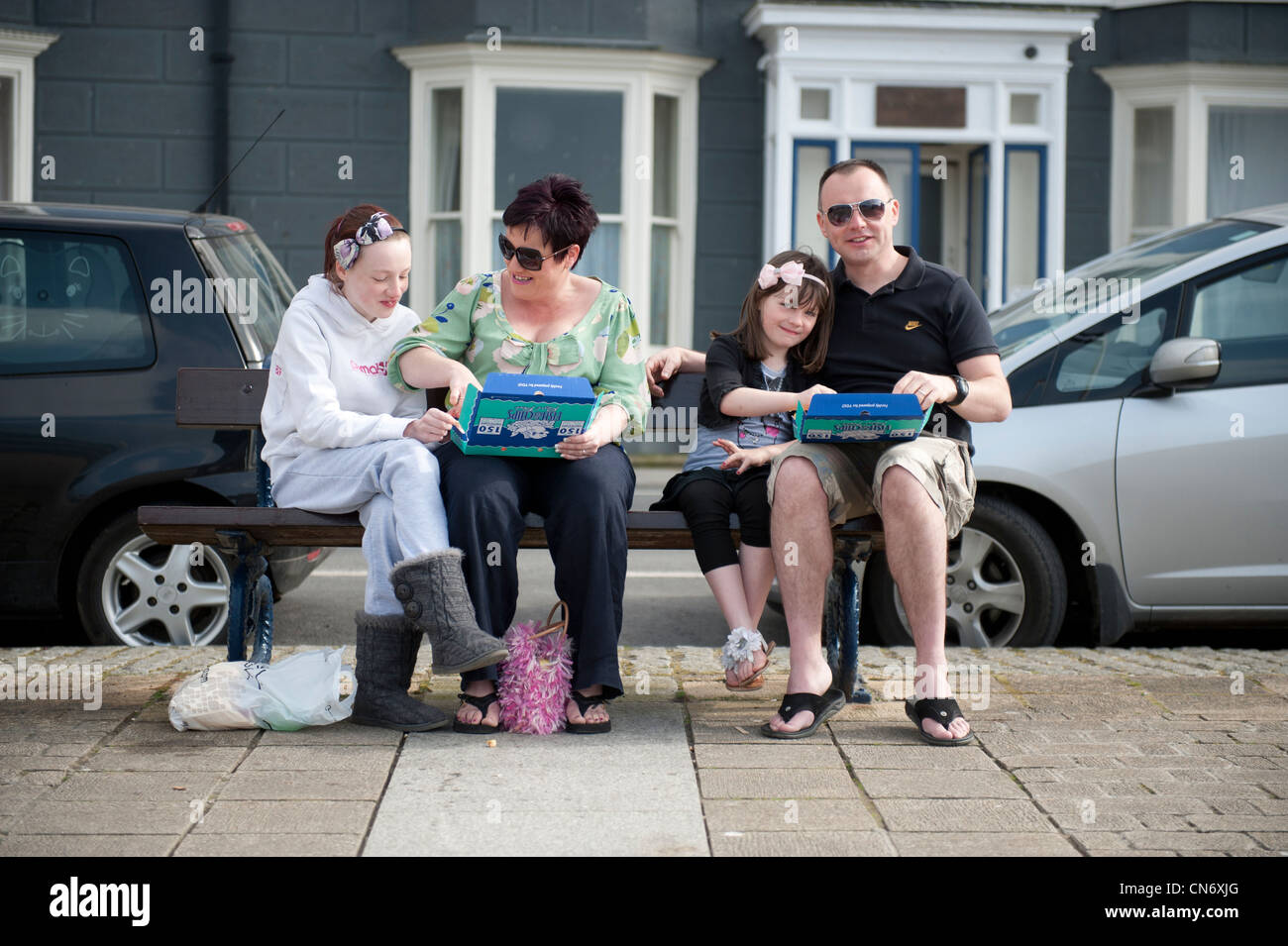 A family sitting on a bench eating take away fish and chips on Aberystwyth promenade, warm spring afternoon, Wales UK Stock Photo