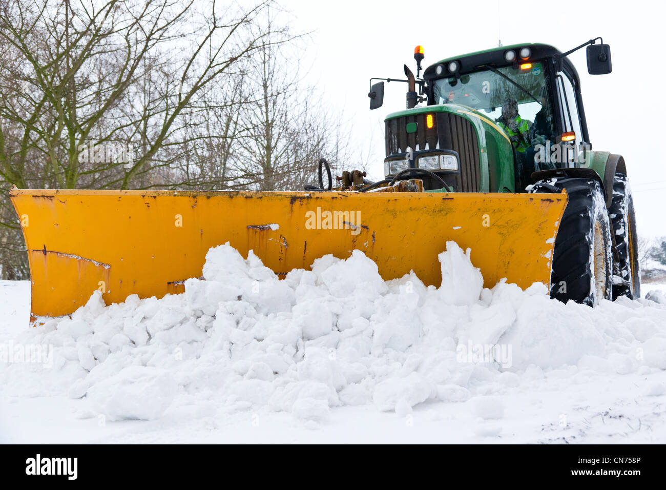 A tractor with a mounted snow plough clearing snow from a country road