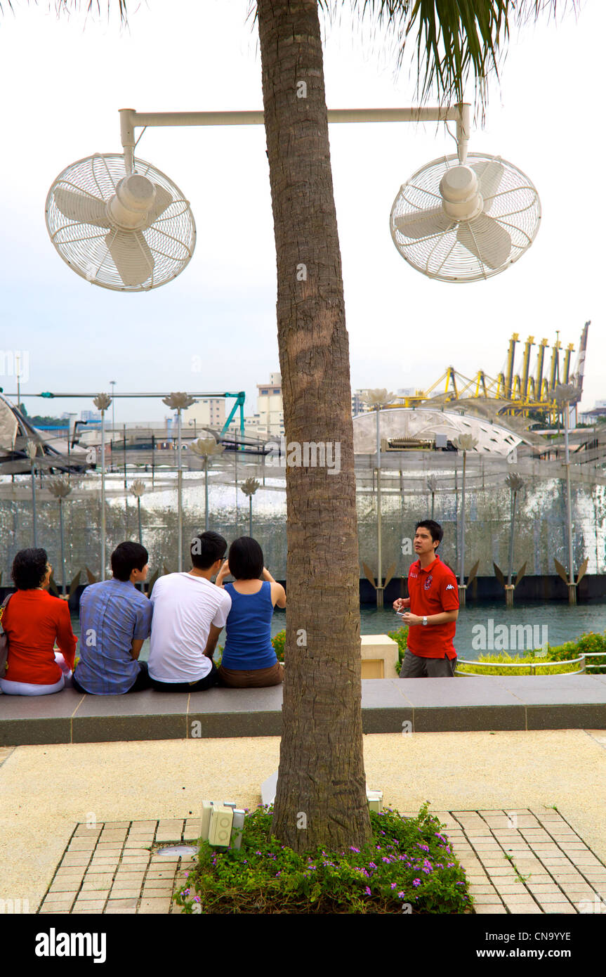 Group of people sitting with their backs to the camera by fans mounted on trees, Sentosa, Singapore Stock Photo