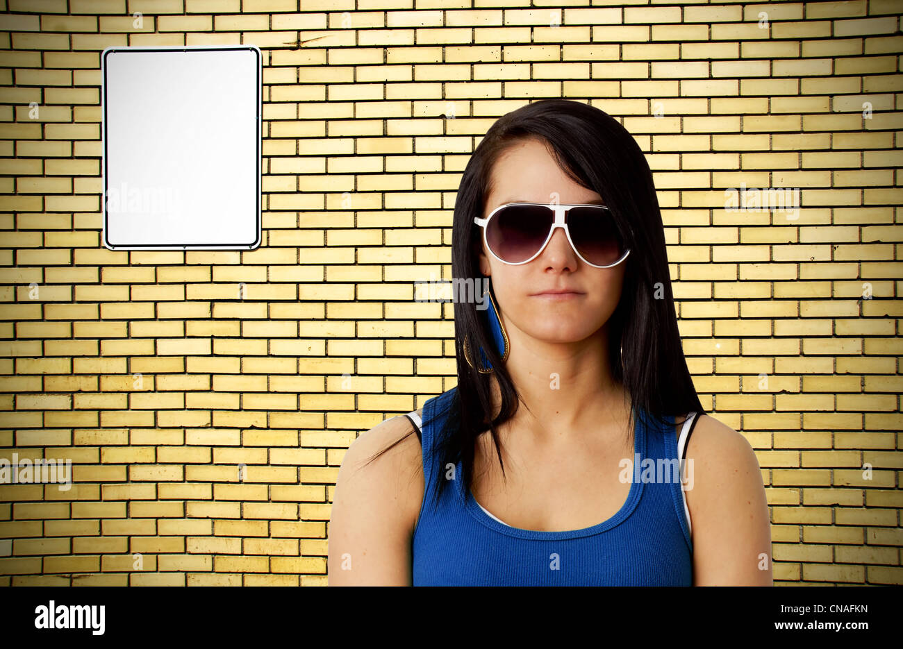 Tough looking young brunette woman with large sunglasses in front of brick wall with blank white sign Stock Photo