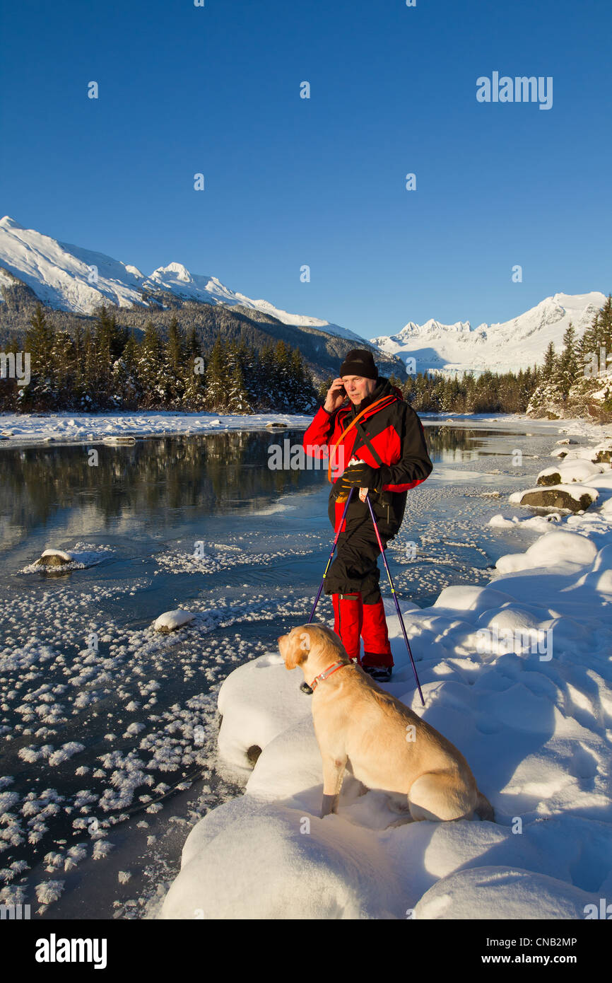 Nordic skier stops to answer his cell phone while his dog sits beside him, Mendenhall River, Juneau, Southeast Alaska, Winter Stock Photo