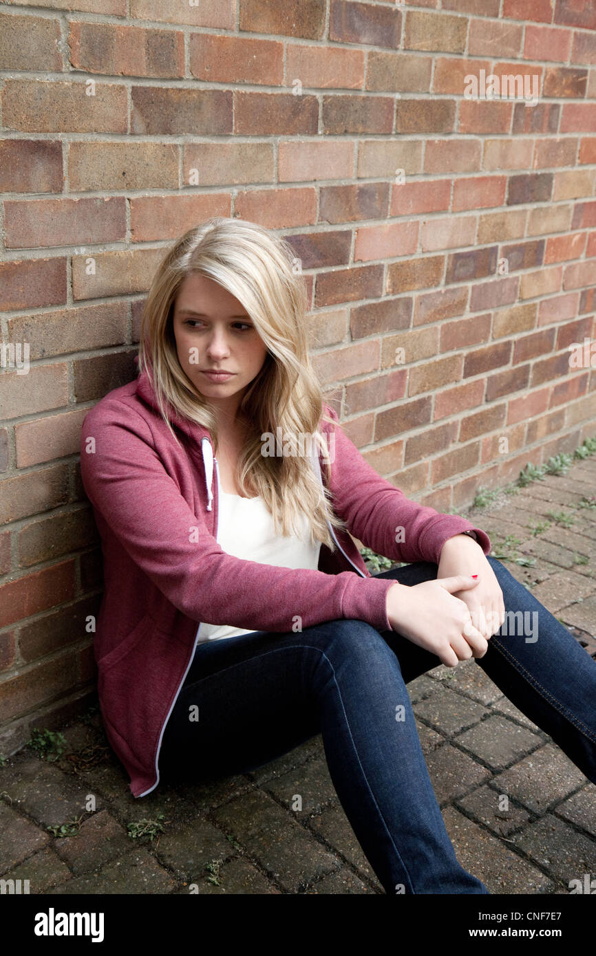 An attractive blonde teenage girl sitting by a brick wall, looking away, Billericay essex UK Stock Photo