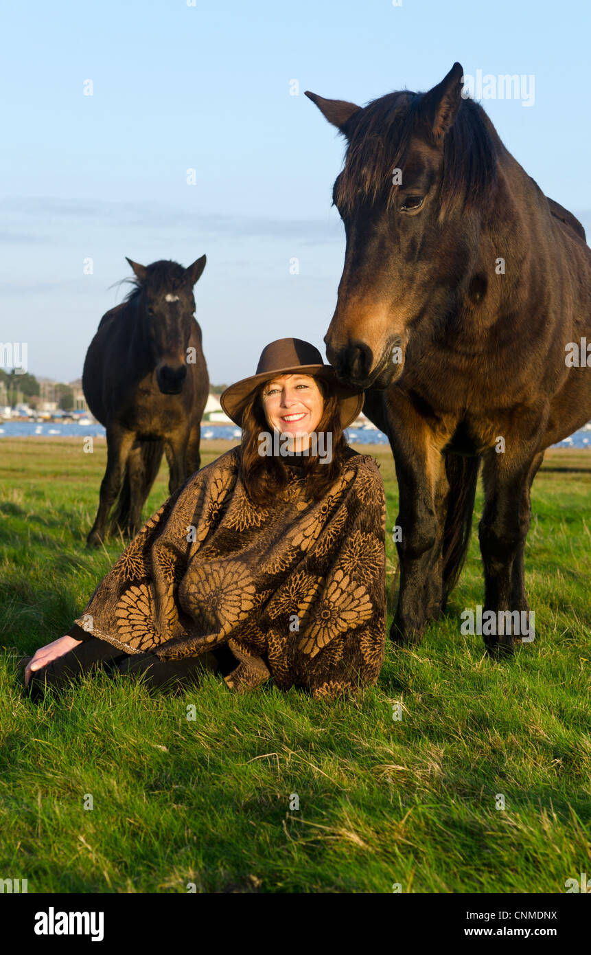 A beautiful woman sitting on the grass wearing a brown poncho wth her horses next to her Stock Photo