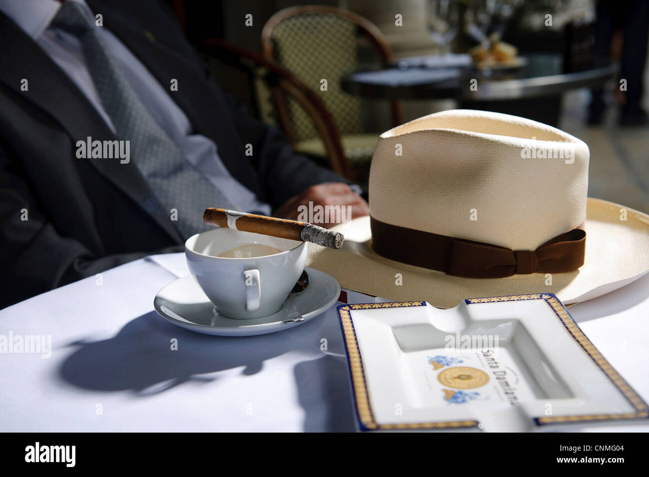 man sitting at outside table at restaurant with trilby hat, ash tray and coffee cup with cigar in sunshine Stock Photo