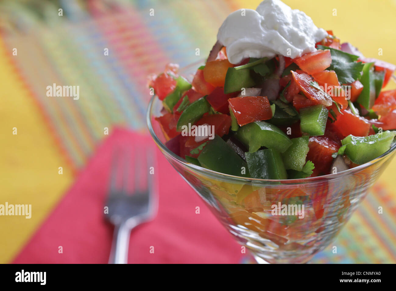 A vegetable salad in a clear glass with a dollop of sour cream, with a fork and napkin on a colorful placemat. Stock Photo