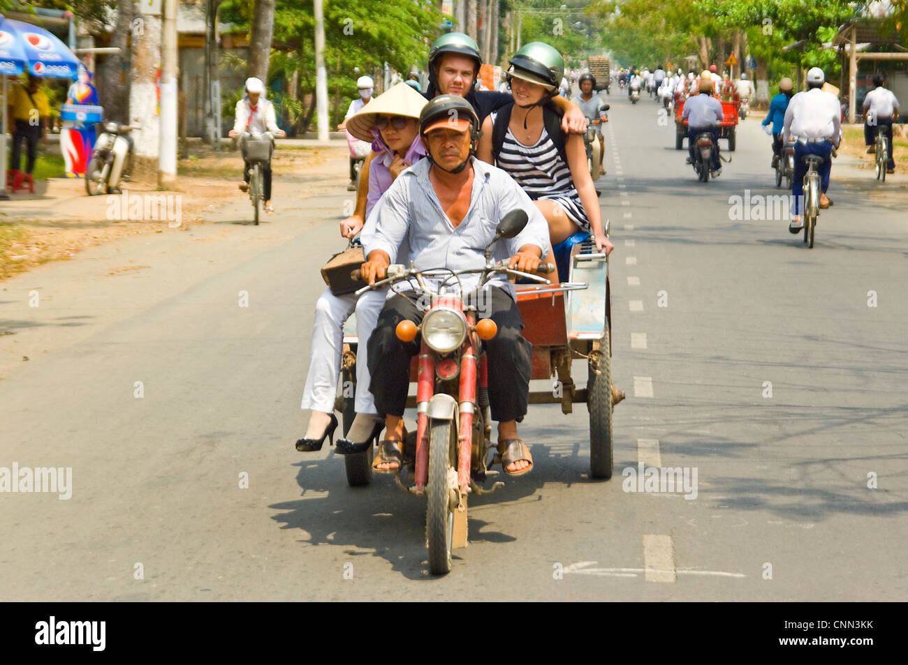 Horizontal portrait of a motorbike and trailer carrying Western tourists and a Vietnamese lady sharing a ride along a busy road. Stock Photo