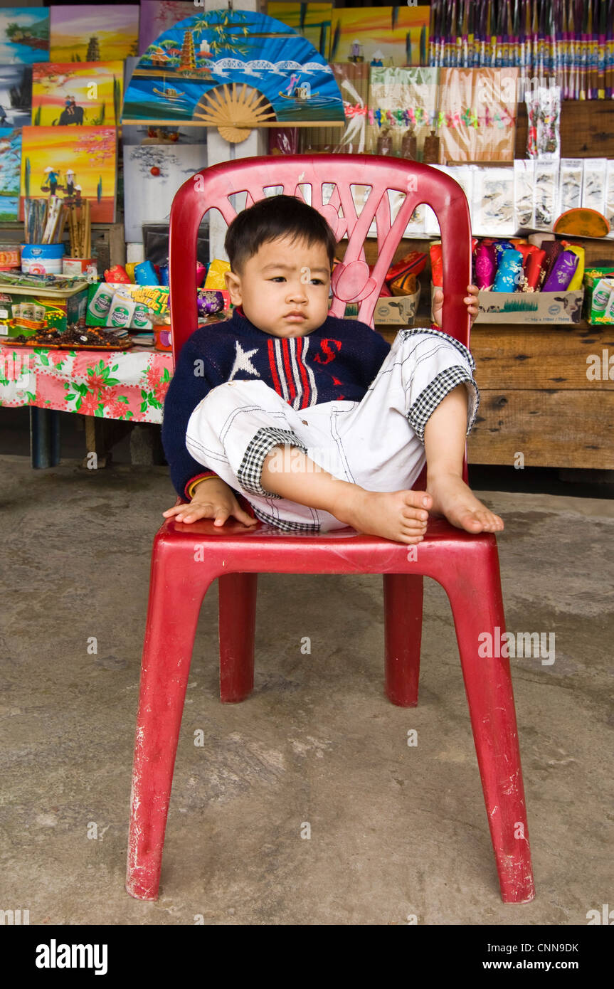 Vertical portrait of a young Vietnamese boy looking bored sitting in a red plastic chair at a market stall. Stock Photo