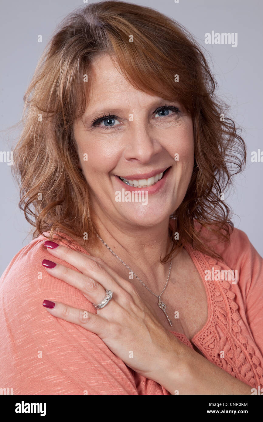 Mature, middle aged, woman in salmon colored shirt, looking at the camera with a happy smile with her left hand on her shoulder Stock Photo