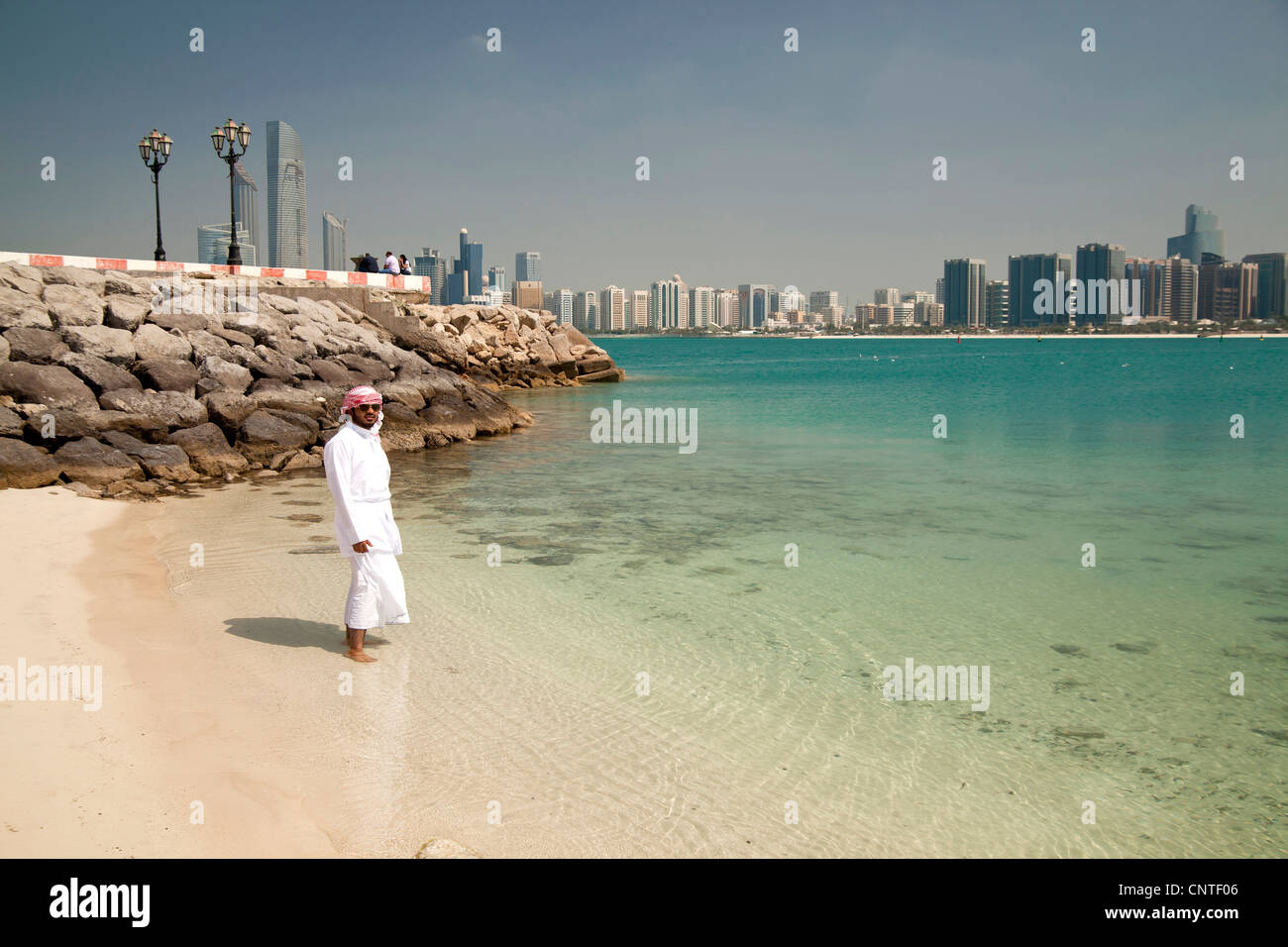 young male local with typical arabic dress Kandora or Dishdasha at the beach in front of the skyline of Abu Dhabi, Stock Photo