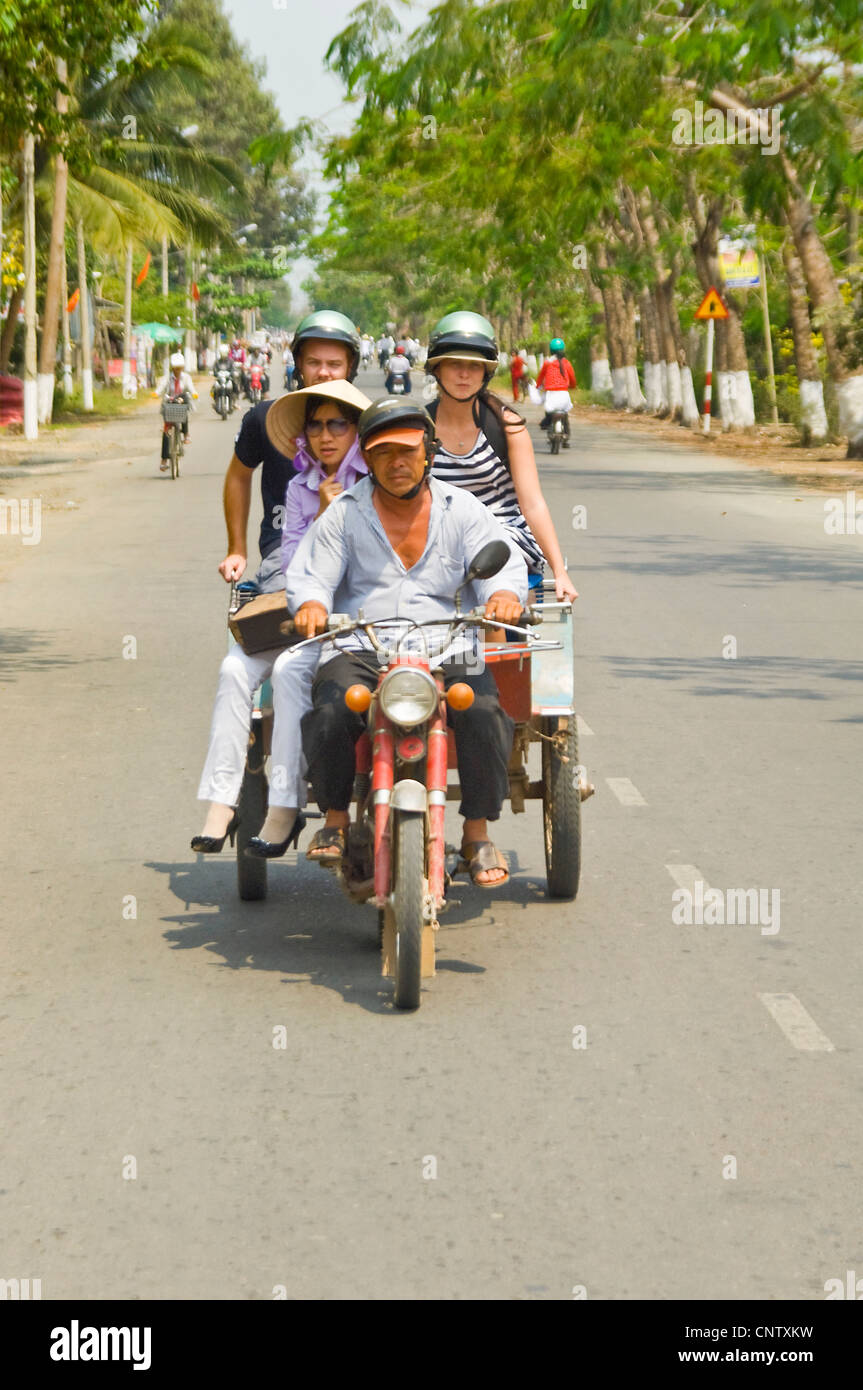 Vertical view of a motorbike and trailer carrying Western tourists and a Vietnamese lady sharing a ride along a busy road. Stock Photo