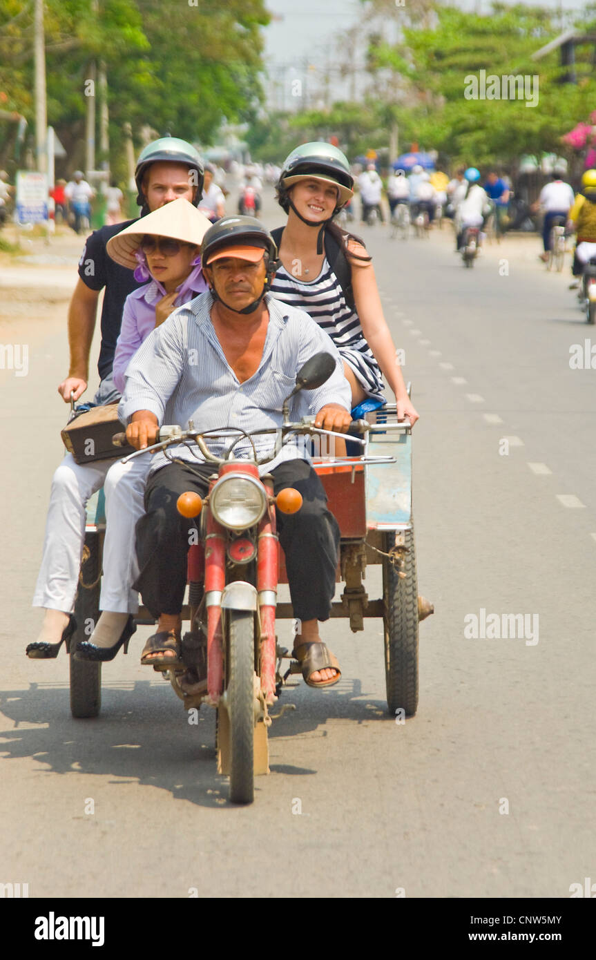 Vertical view of a motorbike and trailer carrying Western tourists and a Vietnamese lady sharing a ride along a busy road. Stock Photo