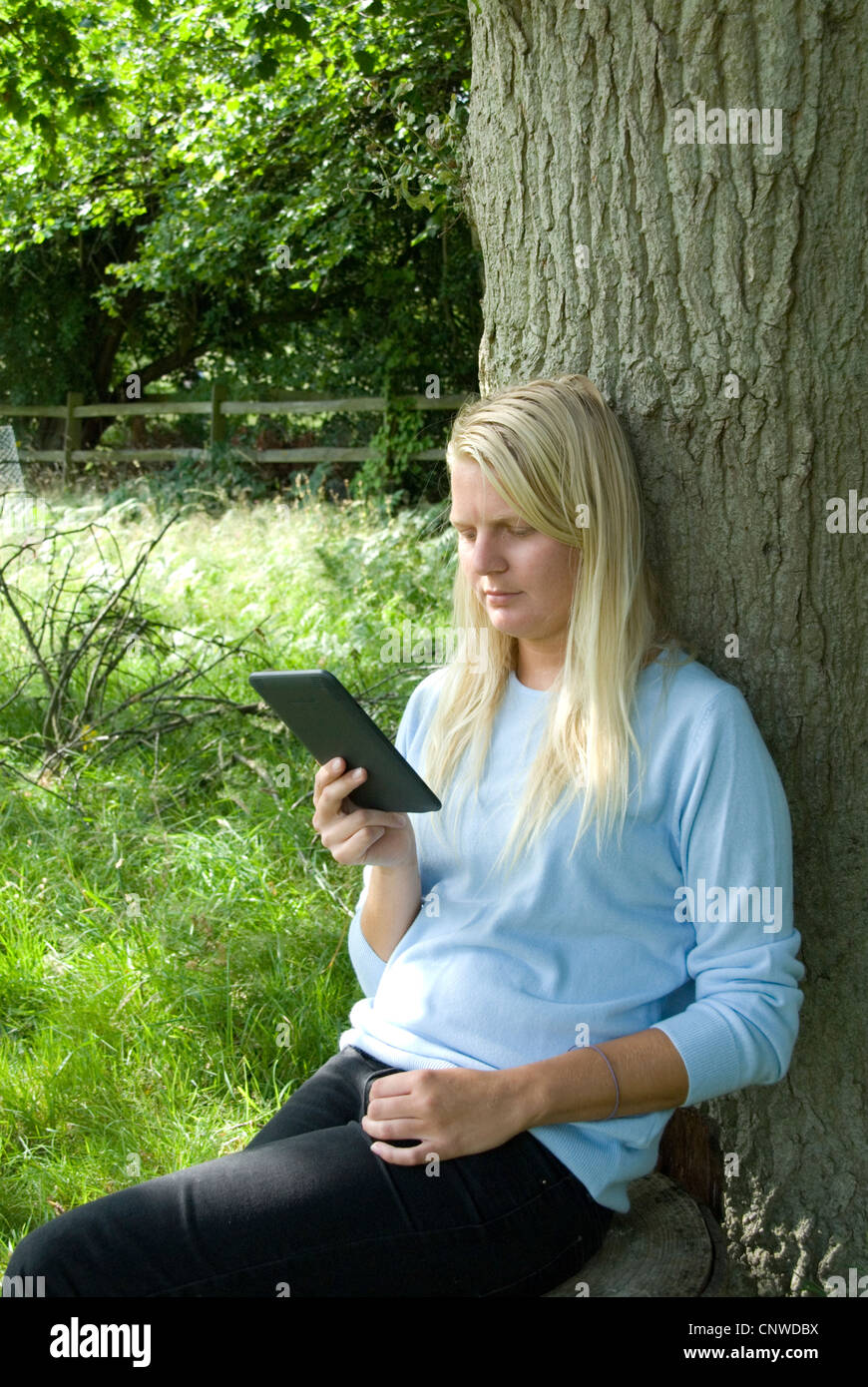 Blonde girl reading an Amazon Kindle electronic e-reader outdoors leaning against a tree Hayley Williamson MR Stock Photo