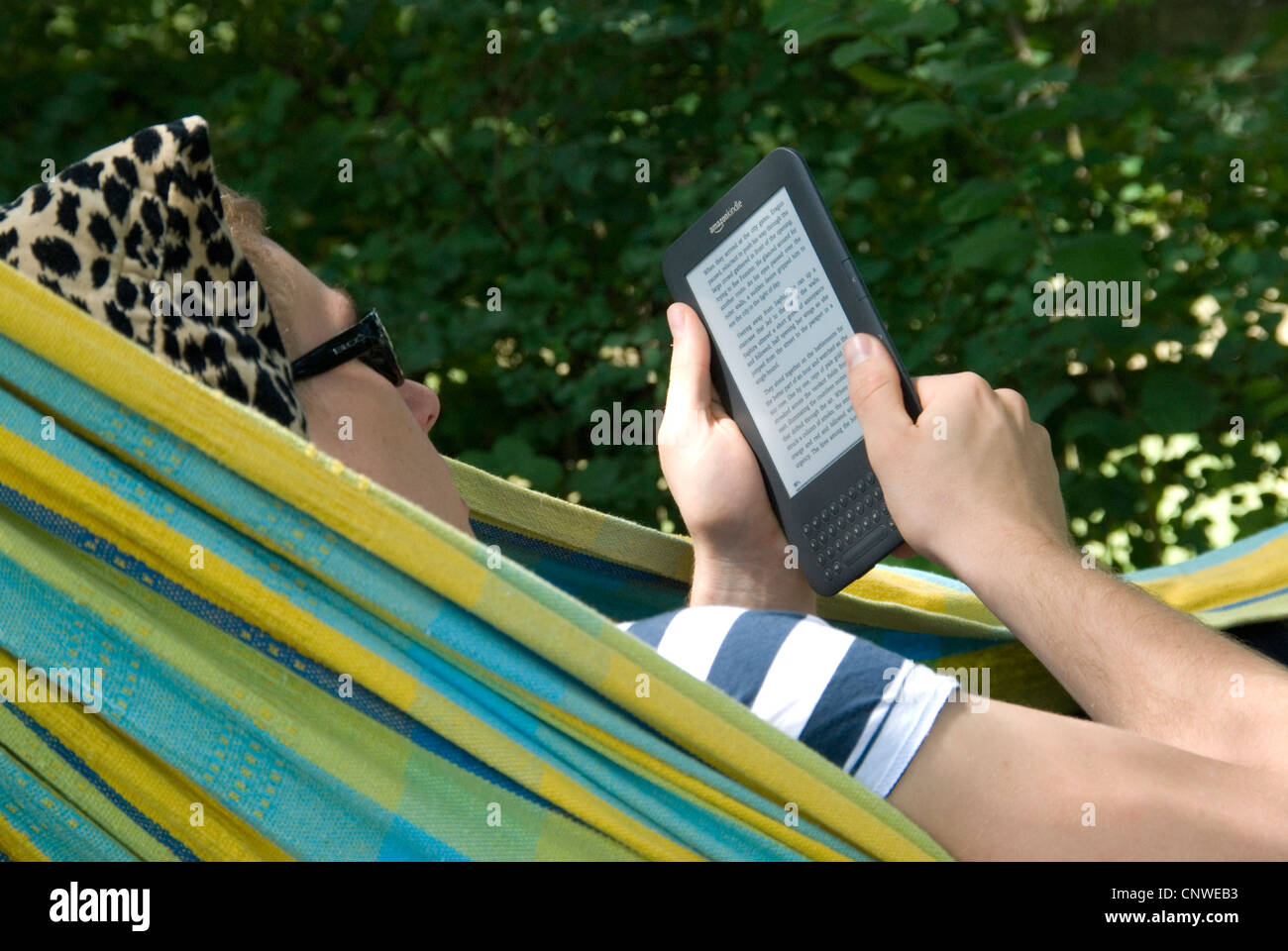 Young man reading Amazon Kindle lying in a hammock outdoors in the garden Luke Hanna MR Stock Photo