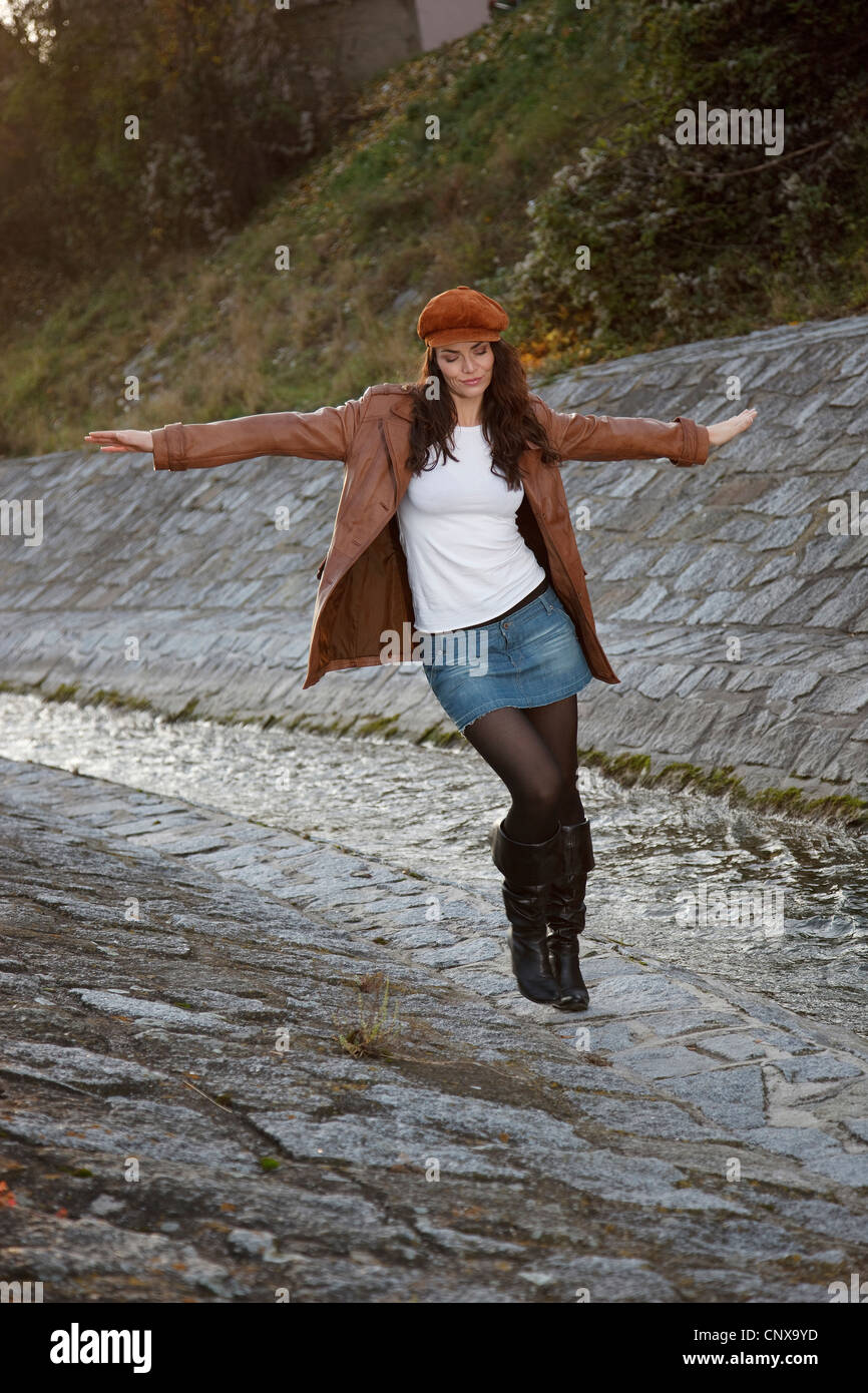 brunette young woman walking with the arms stretched out posing at the edge of a canalised brook in a mini skirt and leather boots Stock Photo