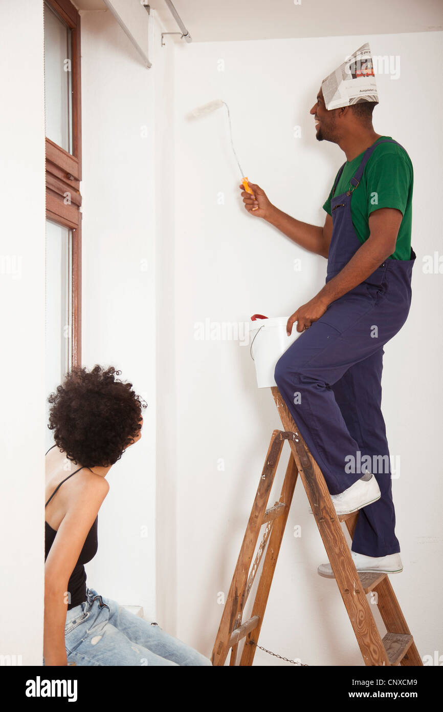 A man standing on a ladder using a paint roller while a woman watches