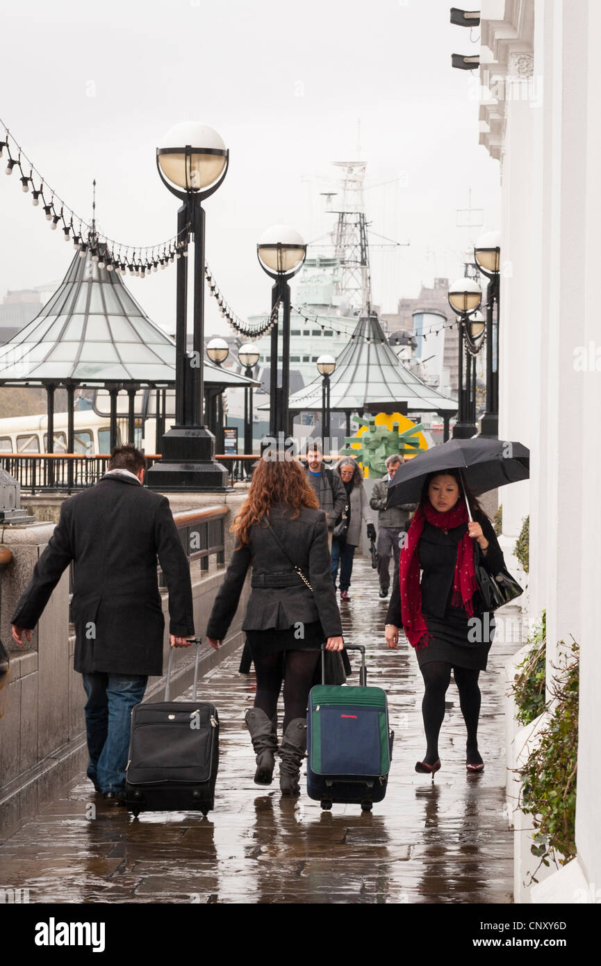 London Bridge Southbank The Queens Walk in rain on grey gray Spring day pedestrians tourists suitcases umbrella Stock Photo