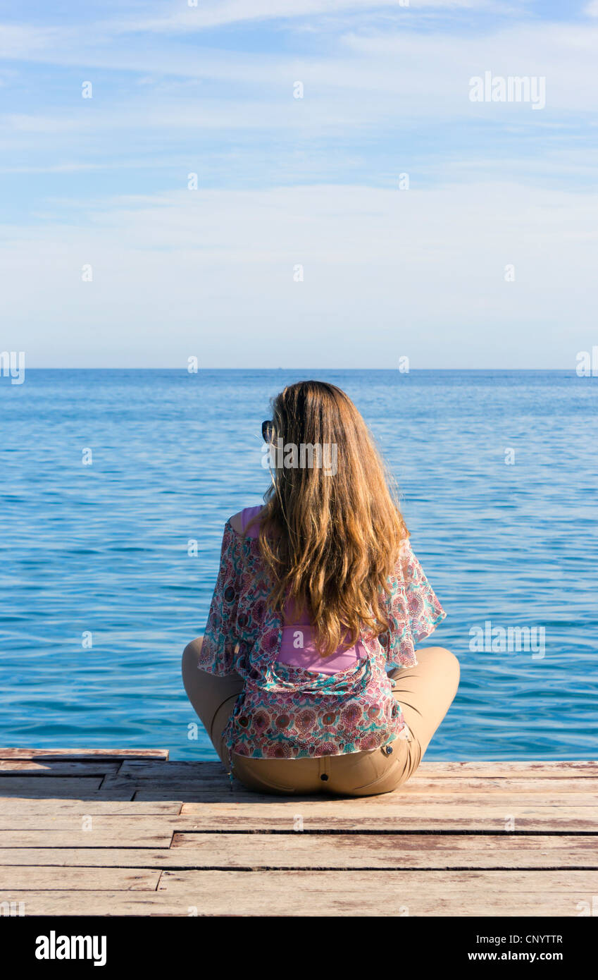 Young woman sat on end of wooden jetty looking out to sea. Stock Photo