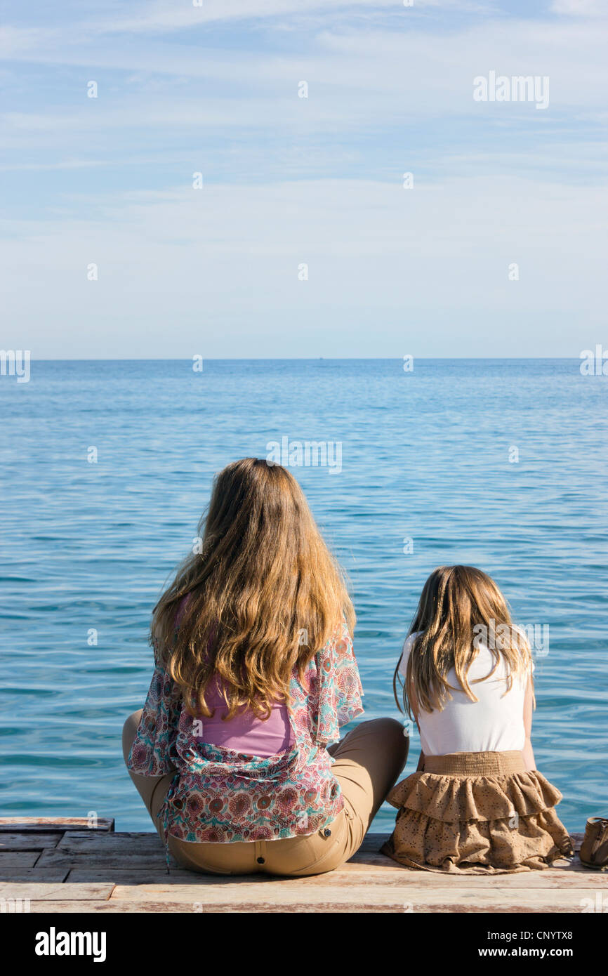 Young woman and little girl sat on end of wooden jetty looking out to sea. Stock Photo