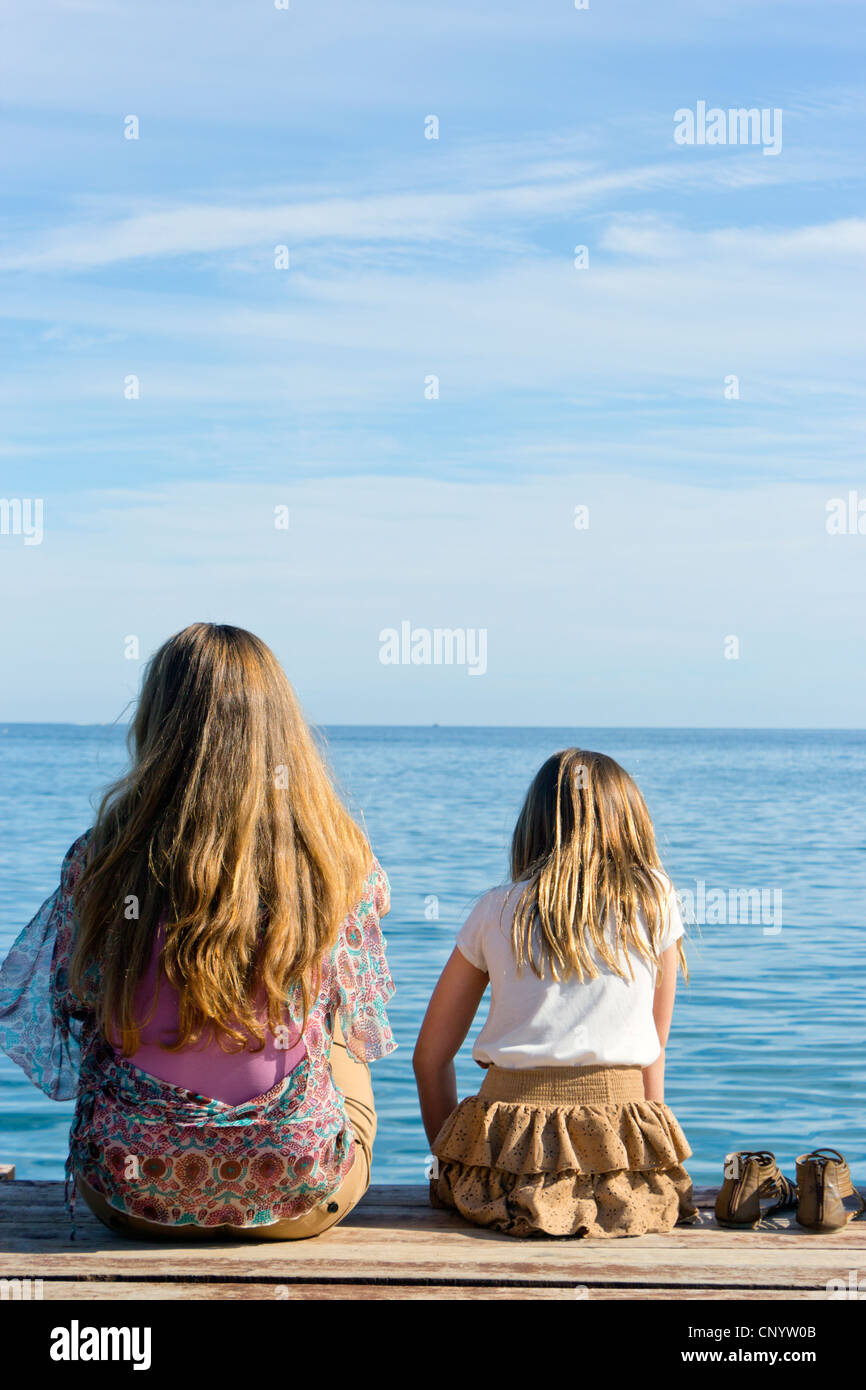 Young woman and little girl sat on end of wooden jetty looking out to sea. Stock Photo