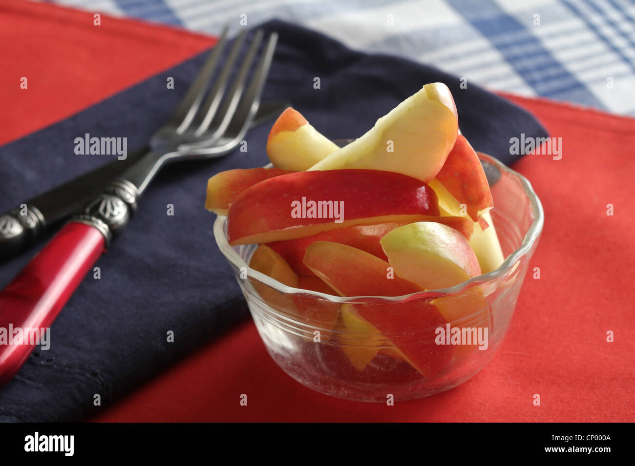 A glass bowl of sliced red apples on a red place mat, with a blue napkin, and a striped blue tablecloth. Stock Photo